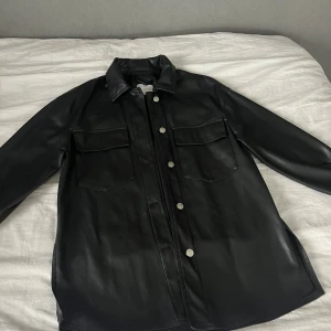Svart Skinnjacka - Stilren svart skinnjacka med klassisk krage, silverfärgade knappar och två stora bröstfickor. Perfekt för dig som vill ha en edgy look med snygga detaljer och lång ärm.