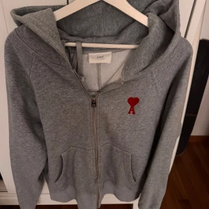 Grå hoodie med röd hjärtlogga från Ami Paris - Grå hoodie från Ami med dragkedja, huva och kängurufickor. Perfekt för chill dagar och enkel att matcha med jeans. Den är knappt använd och därför i väldigt bra skick. Pris går att diskutera och det är bara höra av sig om det finns funderingar.