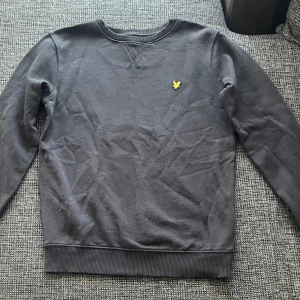 Svart sweatshirt från Lyle & Scott - Svart sweatshirt från Lyle & Scott med klassisk rund hals och ribbade muddar. Tröjan har det ikoniska gula fågel-loggan broderad på bröstet. Perfekt basic att ha i garderoben, passar till allt och är riktigt skön tack vare mjukt bomullsmaterial.