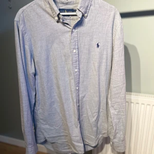 Ljusgrå skjorta från Ralph Lauren - Klassisk ljusgrå skjorta från Ralph Lauren med button-down krage och broderad logga på bröstet. Skjortan har lång ärm, vita knappar och är tillverkad i mjuk bomull. Perfekt för en clean och stilren look.
