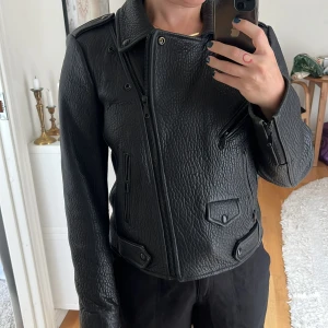 Svart skinnjacka Rebecca Minkoff - Säljer en svart skinnjacka i bikerstil med cool struktur och flera dragkedjor. Jackan har bred krage, axelklaffar och fickor med lock och dragkedja. 