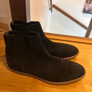 Svarta chelsea boots i mocka - Snygga svarta chelsea boots i mocka med rund tå och platt sula. Skorna har elastiska paneler på sidorna och en diskret söm längs sulkanten. Perfekta för dig som vill ha en stilren och klassisk look.
