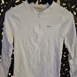 Vit ribbad långärmad tröja amn - Säljer en vit ribbad långärmad tröja från amn i storlek S. Tröjan har en half zip-dragkedja framtill och är tillverkad i ett stretchigt material som sitter snyggt på kroppen. Perfekt till lager-på-lager eller som den är.