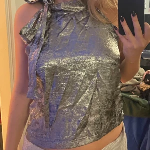 Silvrig halterneck topp med rosett - Supersnygg silvrig halterneck topp med hög krage och en stor rosett vid axeln. Toppen är ärmlös och har en glansig, nästan metallic yta som verkligen sticker ut. Bilder från hemsidan, jag skickar gärna fler om det behövs! Aldrig använd!