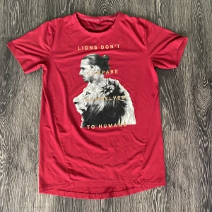 Röd t-shirt med lejontryck - Cool röd t-shirt med tryck av Zlatan och ett lejon på framsidan samt texten 'Lions don't compare themselves to humans'. Perfekt för dig som vill sticka ut med ett unikt motiv. Ny ej använd. 