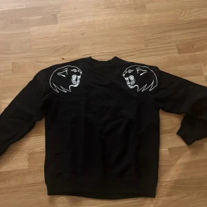 Svart sweatshirt med pantermotiv - Cool svart sweatshirt med två vita pantermotiv på axlarna. Tröjan har en klassisk rund halsringning och långa ärmar. Perfekt för dig som vill sticka ut med ett unikt tryck och streetstyle-vibe.