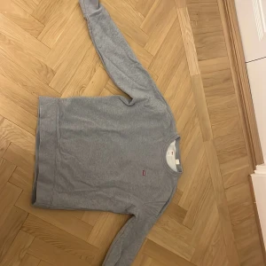 Grå sweatshirt från Levi's i bomull - Säljer en stilren grå sweatshirt från Levi's i mjuk bomull. Tröjan har rund halsringning, lång ärm och en liten röd Levi's-logga på bröstet. Perfekt för en avslappnad look och enkel att matcha med jeans eller joggers.