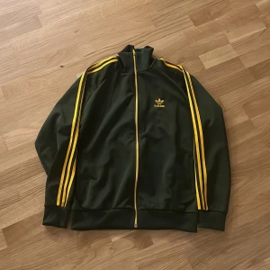 Adidas grön track jacket med gula ränder - Säljer en klassisk grön Adidas track jacket med gula ränder längs ärmarna och gul dragkedja. Tröjan har hög krage, lång ärm och Adidas-logga på bröstet. Perfekt för en sportig och retro look.