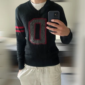 Svart stickad tröja från Diesel | M - Svart stickad tröja från Diesel med stor grå och röd D på bröstet och två röda ränder på ena ärmen. V-ringad modell med ribbade muddar och lång ärm. Perfekt för dig som gillar collegevibbar och vill sticka ut lite extra.