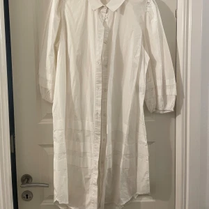 Vit skjortklänning från Zizzi - Stilren vit skjortklänning från Zizzi i bomull med knappar hela vägen framtill. Klänningen har trekvartsärmar med resår vid ärmslut och klassisk krage. Perfekt för en clean och avslappnad look.