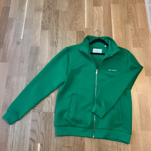 Grön vindjacka från Les Deux - Snygg grön vindjacka från Les Deux med stilren design och diskret logga på bröstet. Jackan har hel dragkedja, ståkrage och två praktiska fickor framtill. Perfekt för dig som gillar cleana och sportiga vibes.