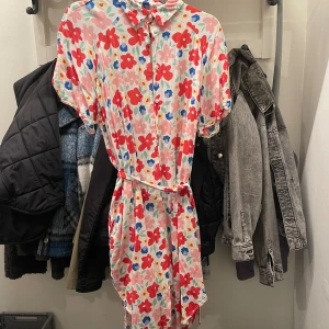 Blommig klänning med bälte - Superfin klänning med färgglada blommor. Klänningen har korta ärmar, krage, knappar framtill och ett matchande bälte i midjan 