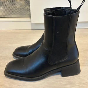 Svarta chelsea boots, stl 38 - Snygga svarta chelsea boots i skinn med fyrkantig tå och låg klack. Klassisk design med resår på sidorna och dragflikar upptill för enkel påtagning. Perfekta för dig som gillar stilrena och tidlösa skor.