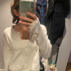 Vit kofta med knappar - Mysig vit kofta med rund hals och knäppning framtill. Tröjan är stickad och har långa ärmar samt ribbade kanter vid hals, ärmslut och nederkant. Perfekt att slänga över ett linne eller t-shirt för en chill look.