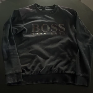 Svart BOSS sweatshirt slim fit L - Svart sweatshirt från Hugo Boss i slim fit med stort BOSS-tryck framtill. Tröjan har rund hals, långa ärmar och ribbade muddar. Perfekt för dig som gillar stilren och enkel design med premiumkänsla. PRISET EJ HUGGET I STEN