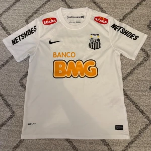 Vit Santos Neymar Jr matchtröja Nike M - Santos FC fotbollströja i vitt med Neymar Jr och nummer 11 på ryggen. Tröjan är tillverkad i Dri-FIT-material och har tryckta sponsorer som BMG, Netshoes och Seara. Klassisk rund hals och klubbmärke på bröstet. Perfekt för dig som älskar brasiliansk fotboll.