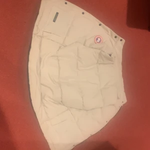 Vest från Canada Goose - Säljer en vest från Canada Goose med klassisk patch på bröstet. Vest är quiltad och har en stilren look med svarta detaljer på insidan. Perfekt för kalla dagar när du vill hålla värmen och ändå se snygg ut.