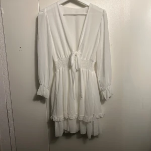 Vit omlottklänning med volangdetaljer - Superfin vit kortklänning med omlottknytning framtill, smockad midja och volangdetaljer vid ärmslut och fåll. Långa ärmar och lätt, flowigt tyg som ger en luftig känsla. Perfekt för dig som gillar romantisk och feminin stil. Endast använd en gång!