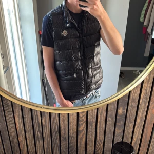 Svart dunväst från Moncler - Snygg svart dunväst från Moncler med klassisk quiltad design och hög krage. Västen har dragkedja framtill, två fickor med dragkedja och Moncler-logga på bröstet. Perfekt att slänga över en hoodie eller t-shirt för en clean look.