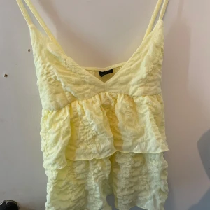 Gul volangtop från Gina Tricot - Supersöt ljusgul linnetopp från Gina Tricot med volanger och smala axelband. Toppen har en v-ringad fram och är tillverkad i ett lätt, krinklat tyg som ger en luftig känsla. Perfekt för varma dagar och somriga outfits. Aldrig använd! 