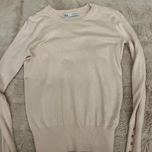 Beige långärmad tröja från Zara - Snygg beige långärmad tröja från Zara med rund halsringning. Tröjan har ribbade muddar och dekorativa guldknappar på ärmsluten som ger en extra touch. Perfekt för lager-på-lager och enkel att matcha med olika outfits.