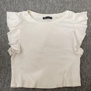 Vit ribbad topp med volangärmar från Zara - Superfin vit ribbad topp från Zara i storlek M. Toppen har korta ärmar med volangdetaljer, den e använd en del.