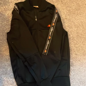 Svart ziptröja från Ellesse XS - Svart långärmad ziptröja från Ellesse i storlek XS. Tröjan har dragkedja framtill, ribbad krage och logoband längs ärmarna. Klassisk sportig look med broderad Ellesse-logga på bröstet. Perfekt för en chill och stilren outfit.