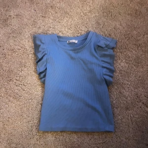 Blå ribbad topp med volangärmar Zara - Supersöt blå ribbad topp från Zara med volangdetaljer på axlarna. Toppen är ärmlös med rund halsringning och har en tight passform. Perfekt för dig som gillar en enkel men ändå trendig look.