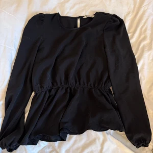 Svart blus med volang från ONLY - Snygg svart blus från ONLY med lång ärm och rund halsringning. Blusen har volang nertill och resår i midjan för en feminin siluett. Stängs med knapp i nacken. Perfekt till jeans eller kjol för en trendig look.