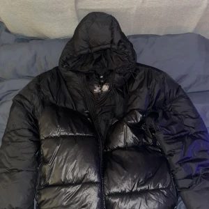 Svart pufferjacka från H&M med huva - Svart pufferjacka från H&M med vattenavvisande och vindtätt material. Jackan har en stor huva, dragkedja framtill och snörning i nederkant för extra skydd mot väder. Perfekt för kalla dagar och har en clean, stilren look, extremt puffy så trots att lappen säger M skulle jag påstå att den passar L eller XL