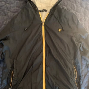 Lyle & Scott vindjacka - Svart vindjacka från Lyle & Scott med gul dragkedja och logga på bröstet. Jackan har huva med snörning, två fickor med dragkedja och grått innerfoder. Perfekt för blåsiga dagar och enkel att matcha med streetwear.