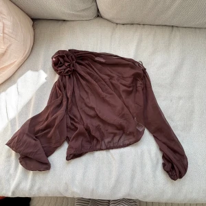 Brun omlottblus med ros från Nelly - Säljer en brun, transparent omlottblus från Nelly med lång ärm och stor dekorativ ros på ena axeln. Blusen har en luftig och flowy känsla, perfekt för dig som gillar unika detaljer och vill sticka ut. Materialet är lätt och ger en snygg drapering.