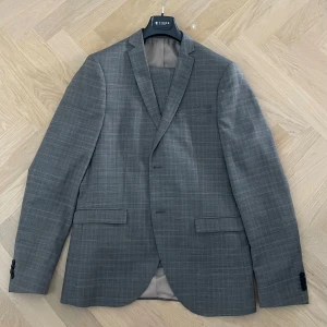 Grå rutig kavaj från Tiger of Sweden - Snygg grå kavaj från Tiger of Sweden med diskret rutigt mönster. Klassisk passform med två knappar framtill, fickor med lock och smal krage. Perfekt för dig som gillar stilrena och moderna plagg. Fodrad insida och stilren design. Storlek 152. Kommer med byxorna 