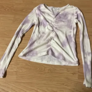 Ljuslila ribbad långärmad topp Kappahl - Säljer en ribbad långärmad topp från Kappahl i vitt och ljuslila med snygg rynkdetalj framtill. Tröjan har ett mjukt, stretchigt material och en smickrande passform. Perfekt för dig som gillar en chill men trendig stil.