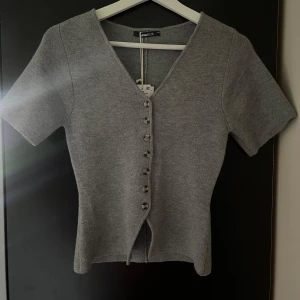 Grå stickad topp från Gina Tricot - Snygg grå stickad topp från Gina Tricot med korta ärmar och v-ringning. Toppen har en rad med dekorativa knappar framtill och är figurnära i modellen. Perfekt att styla med jeans eller kjol för en clean look. Helt oanvänd. 