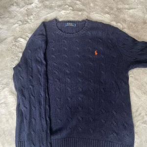 Mörkblå stickad tröja Polo Ralph Lauren - Kabelstickad mörkblå tröja från Polo Ralph Lauren med klassisk rund halsringning och orange broderad logga på bröstet. Tröjan har ribbade muddar och är tillverkad i mjukt material som passar perfekt till lager-på-lager.