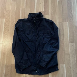 Marinblå safarijacka från gant  - Snygg marinblå safarijacka med hög krage och fyra stora fickor framtill. Jackan har dragkedja och knappar, samt långa ärmar. Perfekt för blåsiga dagar och enkel att matcha med olika outfits. 