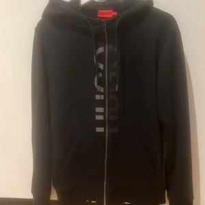 Hugo Boss Zip Hoodie - Svart hoodie från Hugo Boss med stor logga framtill, hel dragkedja och huva med snörning. Tröjan har känguruficka och är långärmad. Perfekt för dig som gillar stilren streetwear med premiumkänsla.