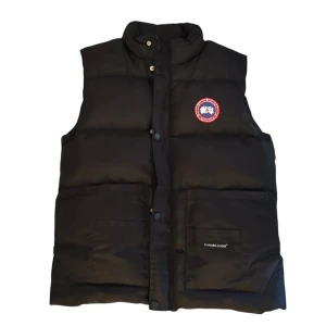 Svart dunväst från Canada Goose - Svart dunväst från Canada Goose med klassisk logga på bröstet och hög krage. Västen har tryckknappar framtill, två stora fickor och quiltad design. Perfekt för lager-på-lager och riktigt snygg till streetwear-stilen. Har ej riktigt bestämt priset så kom med prisförslag!