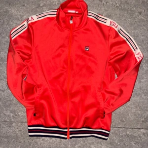 Röd track jacket från Fila -  Kan gå ner i snabbt köp.Röd track jacket från Fila med vita och svarta ränder på axlarna och logga på bröstet. Jackan har hel dragkedja, ståkrage och ribbade muddar med ränder i vitt, rött och marinblått. Perfekt för en sportig och retro look.