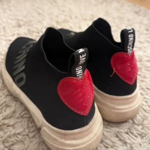Love Moschino svarta sneakers med hjärta - Svarta sneakers från Love Moschino med chunky vit sula och stickad ovandel. På hälen sitter ett rött hjärta i skinn med logga. Texten 'LOVE MOSCHINO' syns på ovansidan och baktill. Snygg och sportig design med platt sula och rund tå.