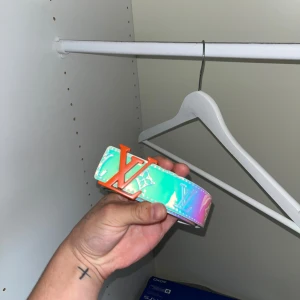 Bälte - Säljer ett bälte med holografisk yta i blå, rosa och turkosa toner. Bältet har en bred design med tydliga monogram och ett stort orange-spänne. Materialet är glansigt skinn och bältet har vita kanter och sömmar. Perfekt statement piece för dig som vill sticka ut.