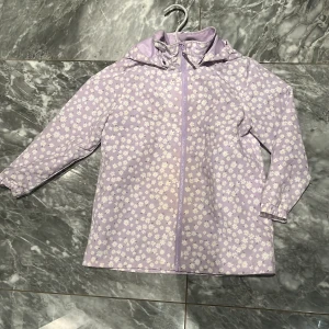 Lila blommig regnjacka från Lindex - Superfin lila regnjacka från Lindex med gulligt blommönster i vitt och gult. Jackan har huva, dragkedja framtill och praktiska fickor med tryckknapp. Perfekt för regniga dagar och har en lätt, smidig känsla. Storlek 128.