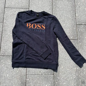 Svart Hugo boss sweatshirt med orange tryck - Svart sweatshirt från Hugo boss med stort orange logotryck på bröstet och blå siffror under. Klassisk rund hals och långa ärmar. Tillverkad i mjuk bomull som är skön mot huden. Perfekt för en clean och stilren look.