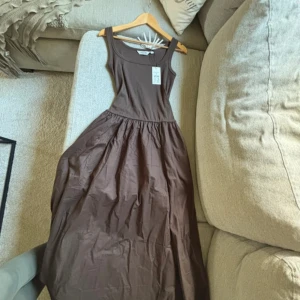 Brun långklänning från Kappahl - Snygg brun långklänning från Kappahl med breda axelband och rundad halsringning. Klänningen har en figurnära överdel och en utställd kjol med fickor i sidorna. 