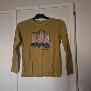 Gul långärmad tröja med surikat-tryck - Gul långärmad tröja med ett roligt tryck av tre surikater som DJ:s och texten 'DJ CREW IN DA HOUSE' på bröstet. Tröjan har rund hals och är gjord i mjuk bomull, perfekt för dig som gillar unika och sköna plagg med humor.