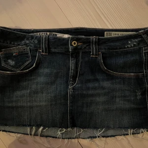 Mini jeanskjol från Tommy Hilfiger - Snygg mörkblå minijeanskjol från Hilfiger Denim med rå kant nertill. Kjolen har slitna detaljer och coola kontrastsömmar. Insidan har ett unikt tryck med amerikanska flaggan. Midjemått 45 cm rakt över i höjd med knappen.