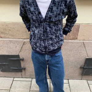 Mörkblå kofta med grafiskt mönster - Säljer en mörkblå kofta med vitt grafiskt mönster och knappar framtill. Koftan har v-ringning, ribbade muddar och lång ärm. Perfekt för dig som gillar retrostil och vill sticka ut lite extra.