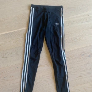 Svarta Adidas leggings med vita ränder - Svarta leggings från Adidas med klassiska vita ränder längs sidorna och Adidas-logga på höften. Tight passform och stretchigt material som sitter snyggt. Perfekta för en sportig och avslappnad look.
