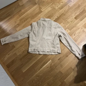 Beige jeansjacka från 157, storlek L - Snygg beige jeansjacka från 157 i klassisk modell med knappar framtill och två bröstfickor. Jackan har raka linjer, lång ärm och justerbara knappar i midjan. Perfekt för dig som gillar en clean och stilren look.
